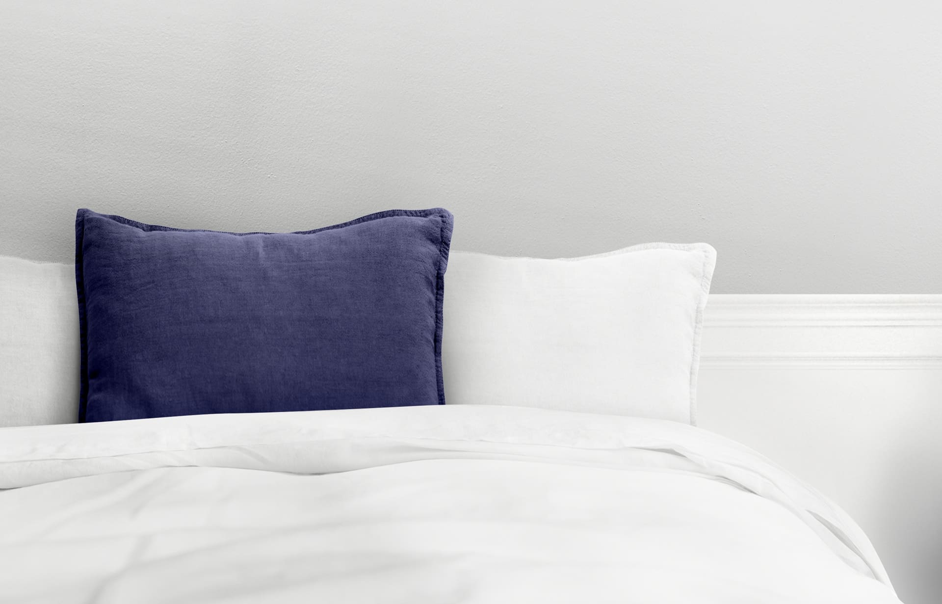 A neatly made bed with white bedding, two white pillows, and one dark blue pillow against a light gray wall—perfectly fresh thanks to local Comforter Cleaning Service.