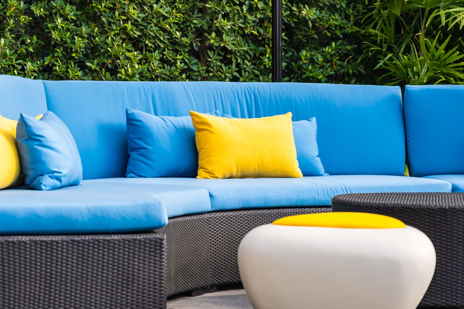 Outdoor wicker sofa with blue cushions and yellow throw pillows, next to a round white ottoman with a yellow top, set against a leafy green hedge.