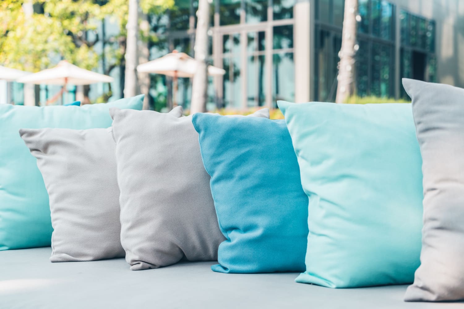A row of light blue and gray cushions arranged on outdoor seating, with a blurred background of greenery and glass windows.