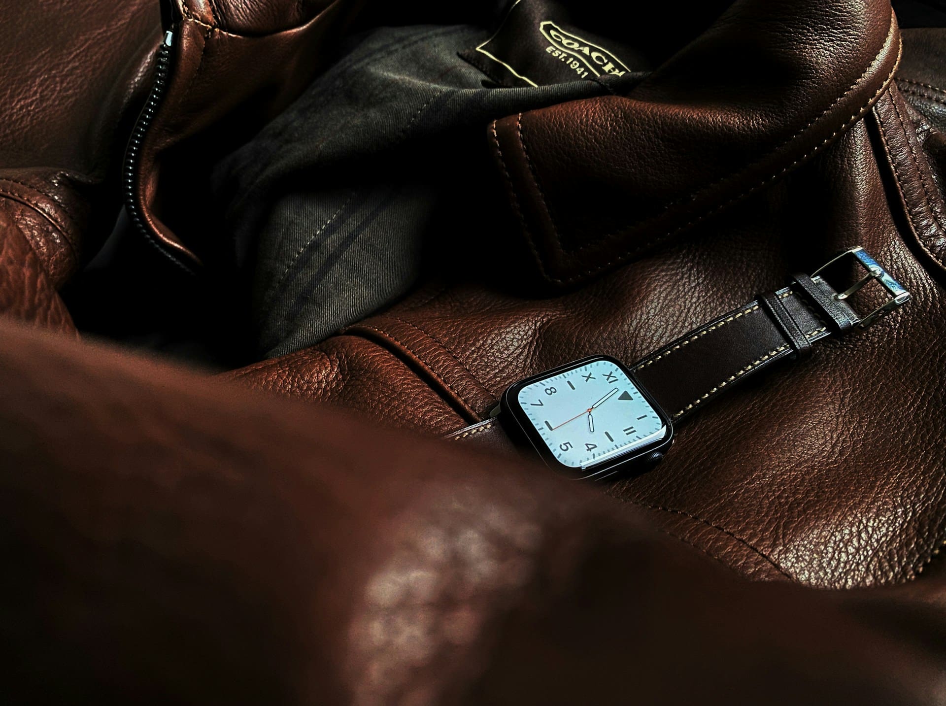 A smartwatch with a brown leather band rests on a brown leather jacket with a visible Coach brand label.
