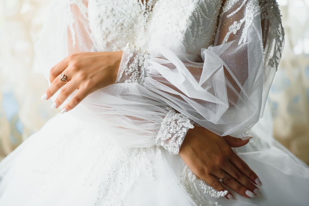 Close-up of a person wearing a white, beaded wedding dress with sheer, puffed sleeves, showing hands with a ring and manicured nails resting on the dress.