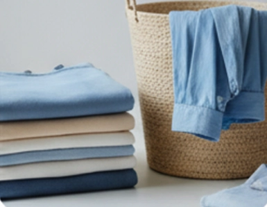 A stack of neatly folded clothes sits next to a woven basket holding a light blue shirt draped over its edge.
