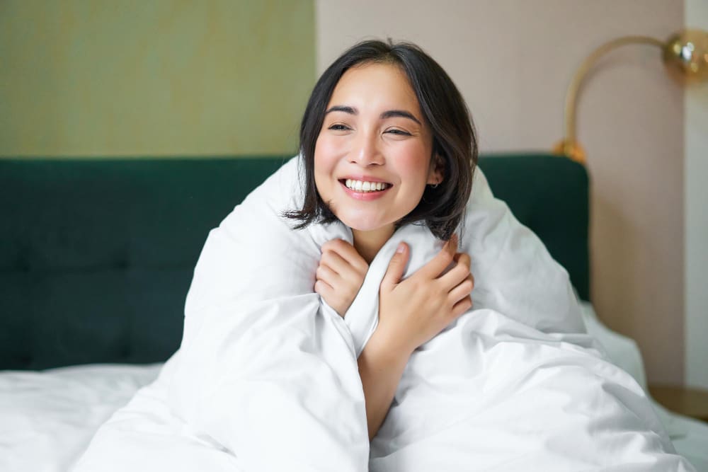 A young woman sitting on a bed, wrapped in a white blanket, smiling and looking slightly off camera in a brightly lit room.