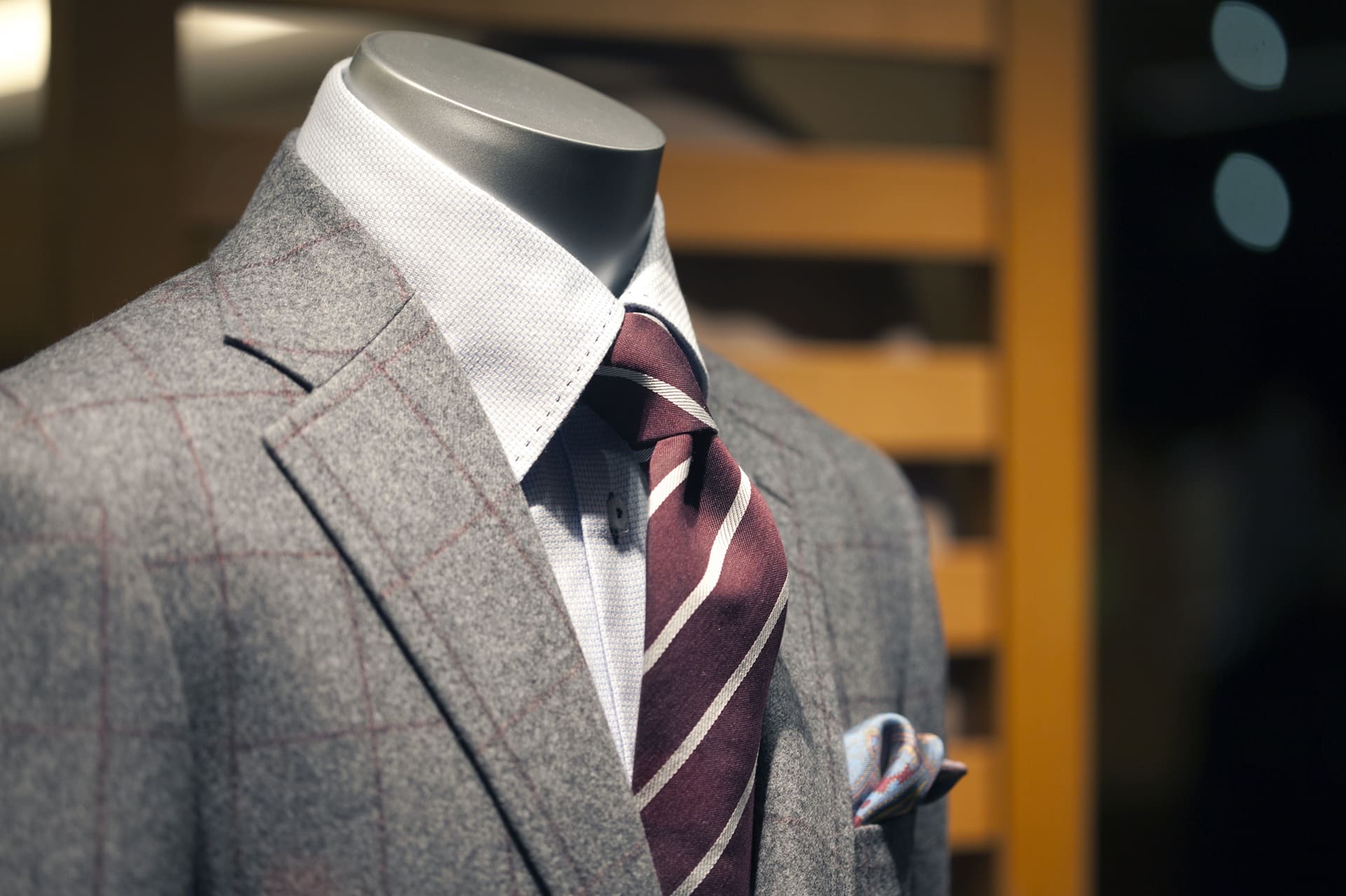 A close-up of a mannequin dressed in a grey plaid suit, white dress shirt, and striped maroon tie, with a pocket square visible in the breast pocket.