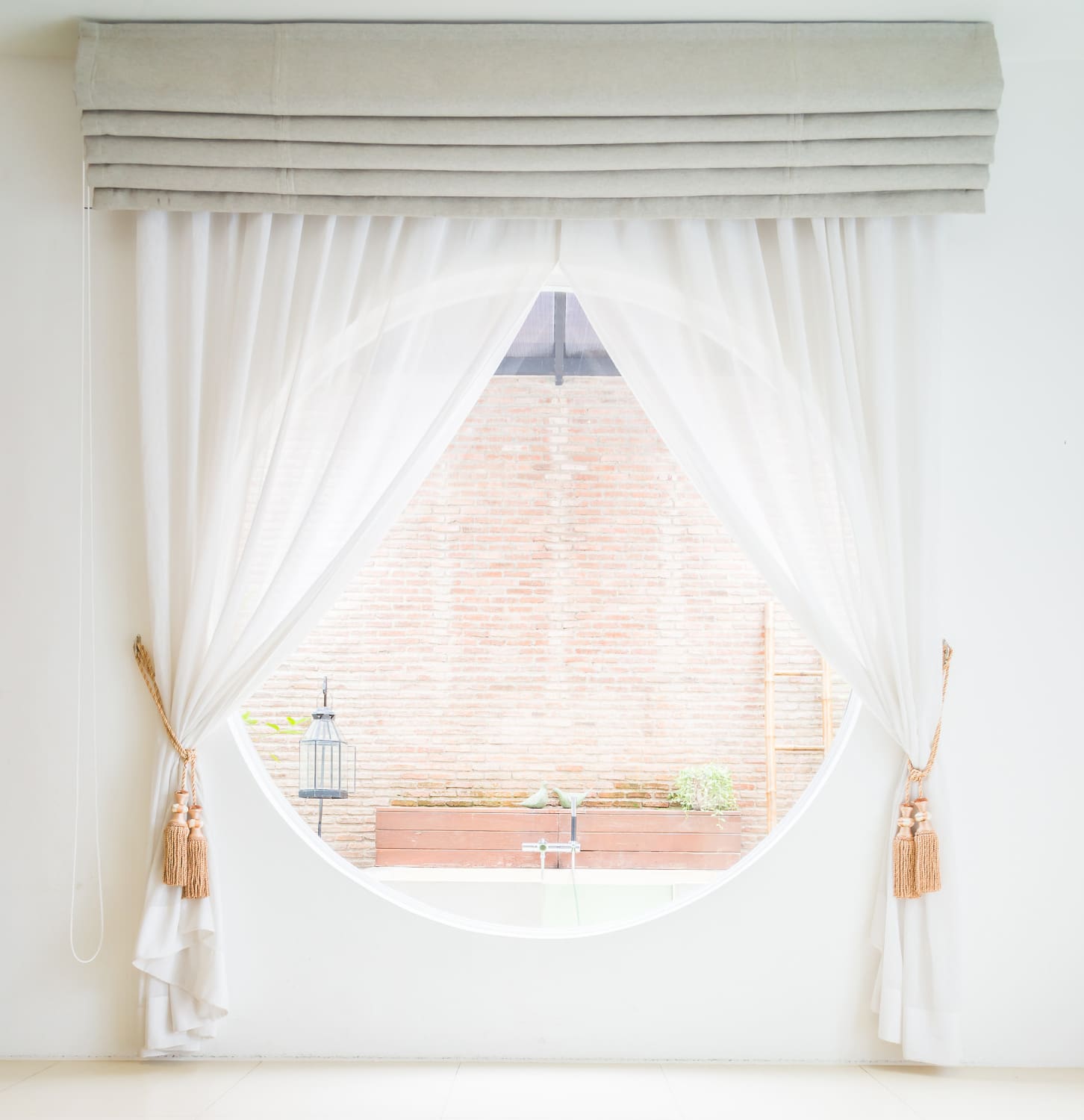 White curtains with tassels are drawn aside, framing a large round window that looks out onto a brick wall and a small garden area with potted plants.