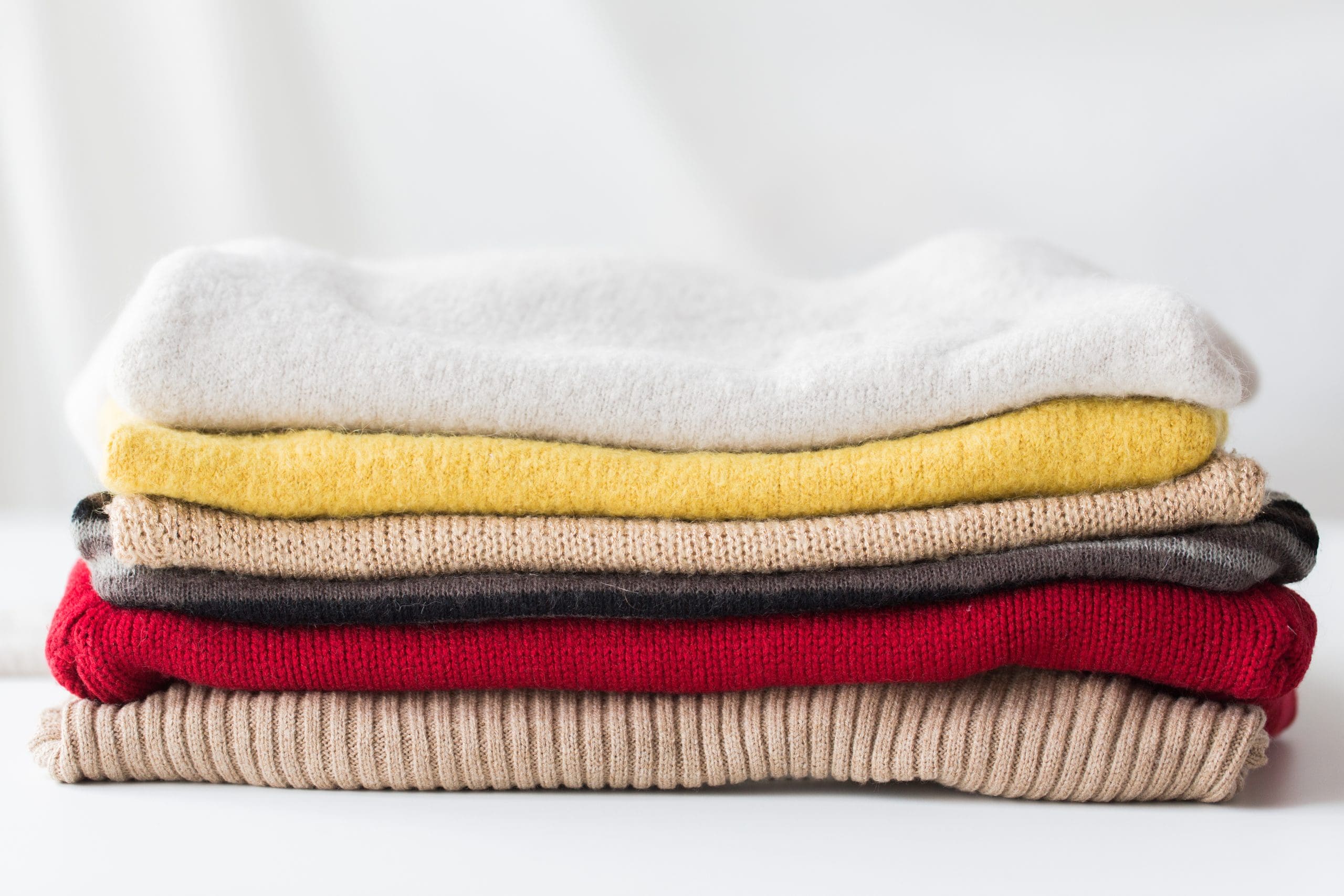 A neatly folded stack of sweaters in various colors including white, yellow, beige, gray, red, and brown on a white background.