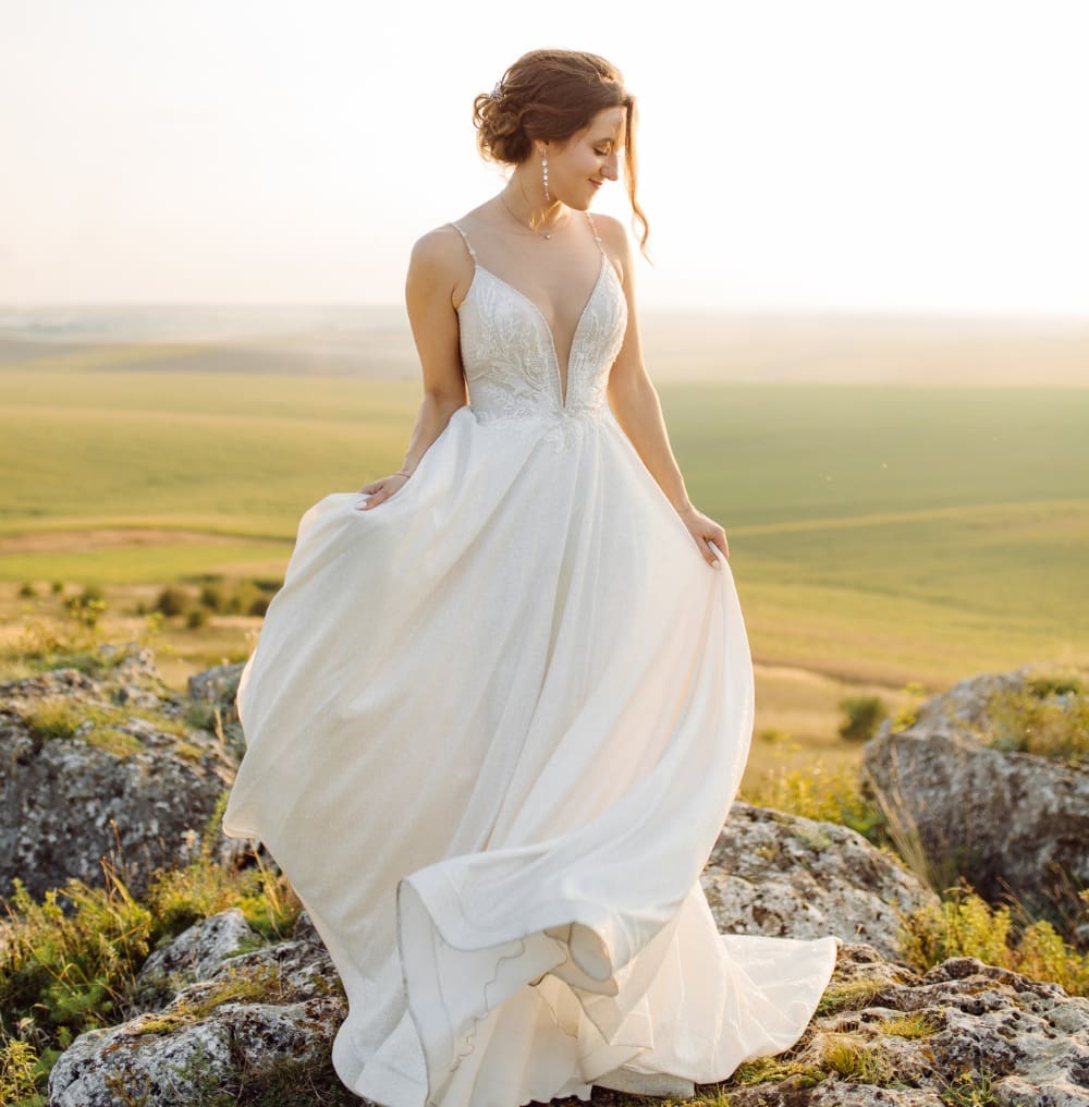 Woman in a white sleeveless wedding dress standing outdoors on rocky ground with a green field and clear sky in the background, holding the sides of her dress.