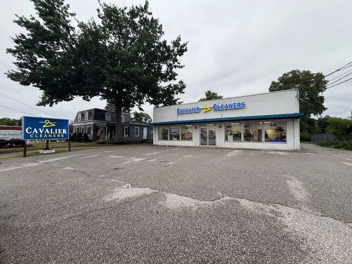 A dry cleaning business named Cavalier Cleaners with a large sign and a white building sits next to a tree and house, with an empty parking lot in front.