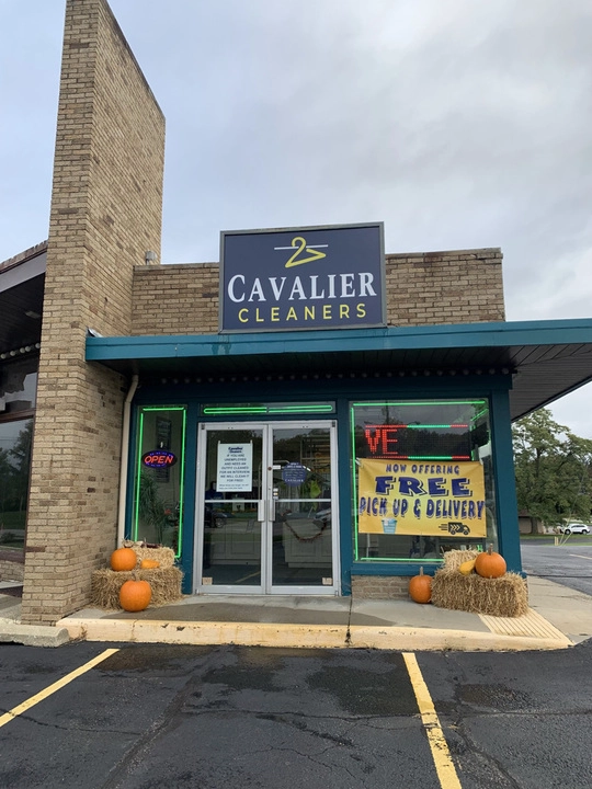 Storefront of Cavalier Cleaners with pumpkins and hay bales outside, an "OPEN" sign, and a window sign advertising free pick up and delivery services.