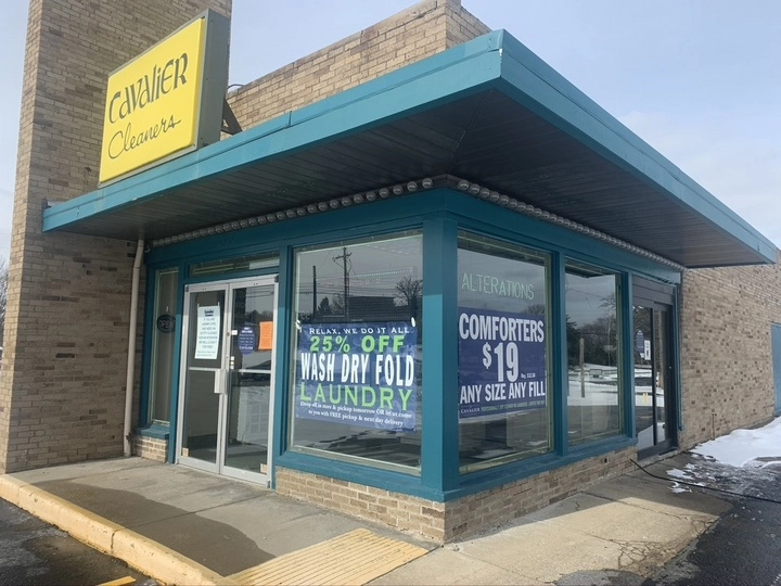 Corner view of a dry cleaners shop with signs advertising 25% off laundry services and $19 comforter cleaning; the building is brick with teal trim and large windows.