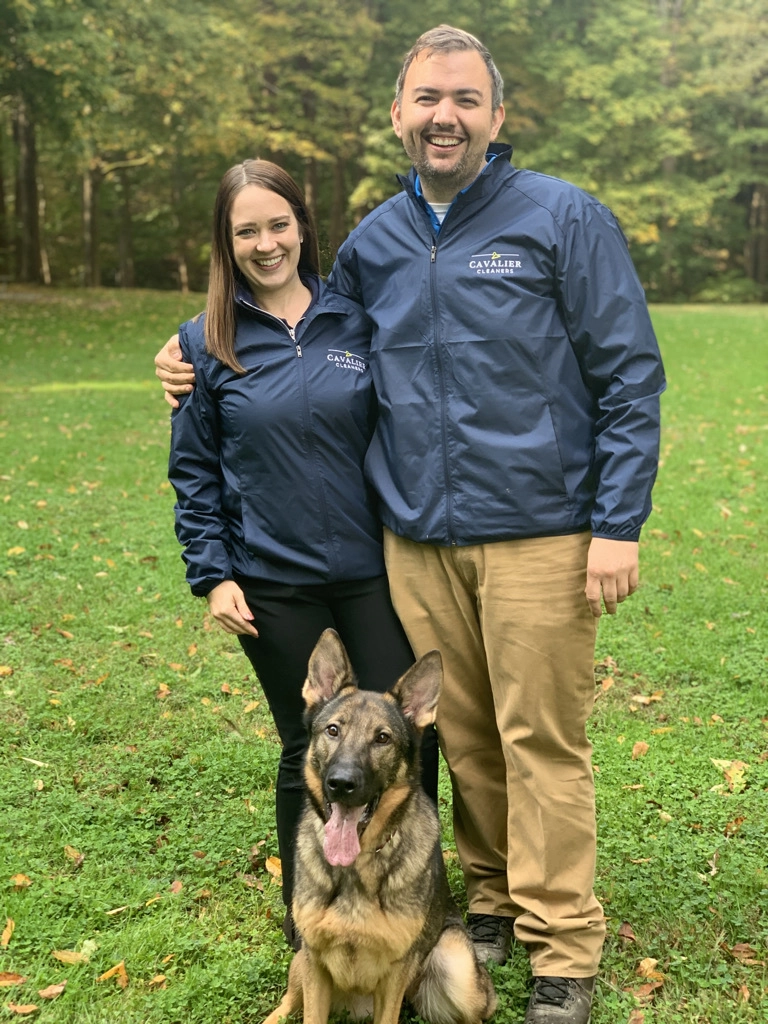 A man and woman in matching blue jackets stand outside on grass with a German Shepherd sitting in front of them; trees are in the background.