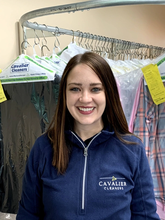 A woman wearing a "Cavalier Cleaners" jacket stands in front of a rack of dry-cleaned clothes inside a dry cleaning business.