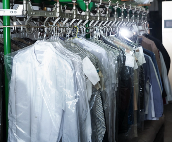 Clothes on hangers covered in plastic, arranged on a conveyor rack at a dry cleaning facility.