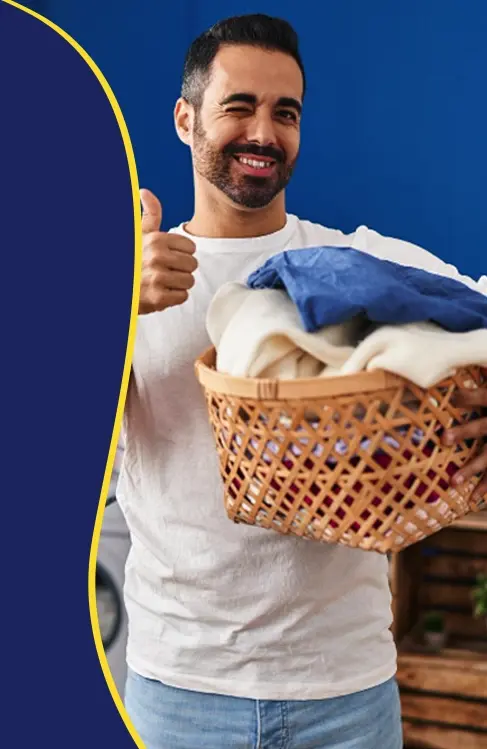 A man holding a laundry basket filled with clothes gives a thumbs up and winks, standing indoors against a blue background.