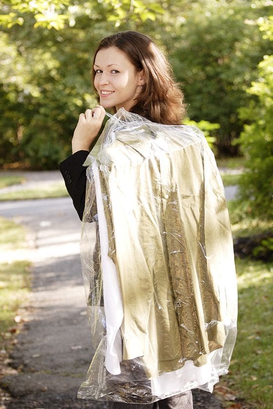 A woman outdoors holds a hanger with clothes covered in a plastic dry cleaning bag, looking back over her shoulder and smiling.