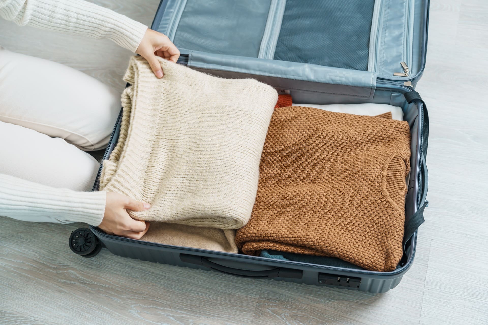 Person neatly packing folded sweaters into an open suitcase on a light wooden floor.