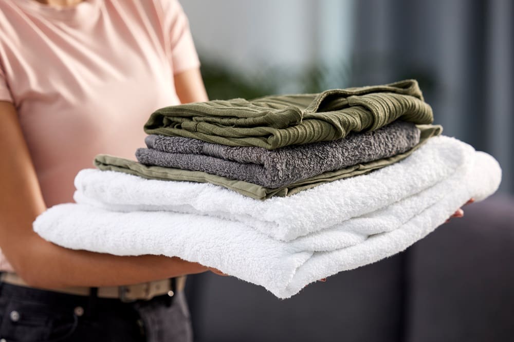 A person holding a stack of neatly folded towels and clothes, including white towels and green and gray garments.
