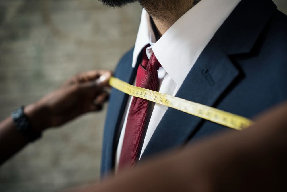 A person in a suit and red tie is being measured across the chest with a yellow measuring tape by another individual.