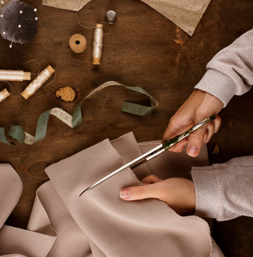 Hands cutting beige fabric with scissors on a wooden table, surrounded by spools of thread, a thimble, a measuring tape, and sewing supplies.