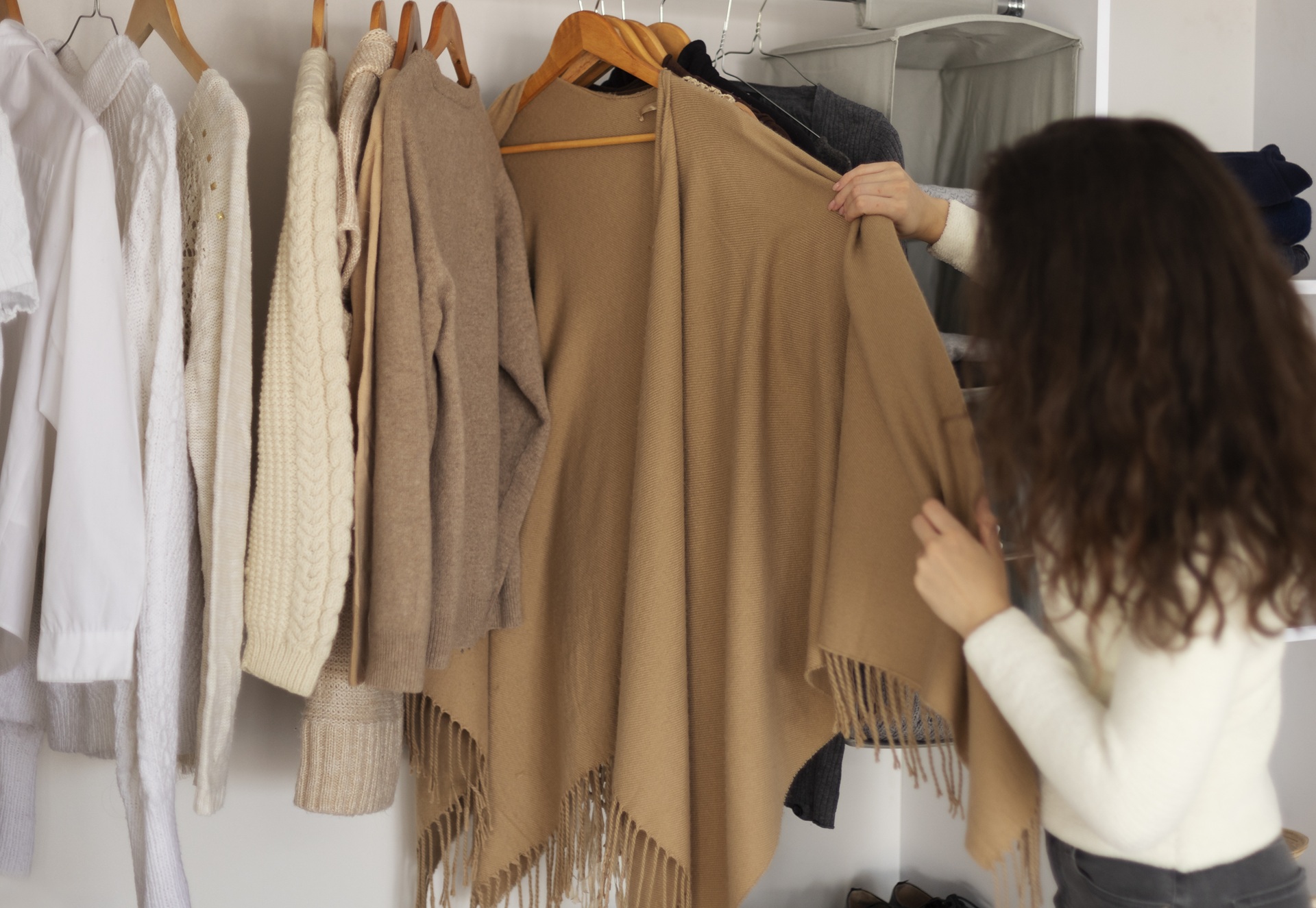 A person stands in front of a wardrobe, holding a beige fringed shawl; various sweaters and shirts hang beside it.