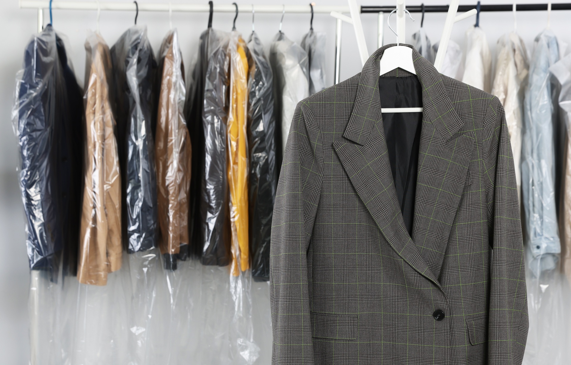 A gray plaid blazer on a hanger is displayed in front of a clothing rack with various garments covered in plastic dry-cleaning bags.
