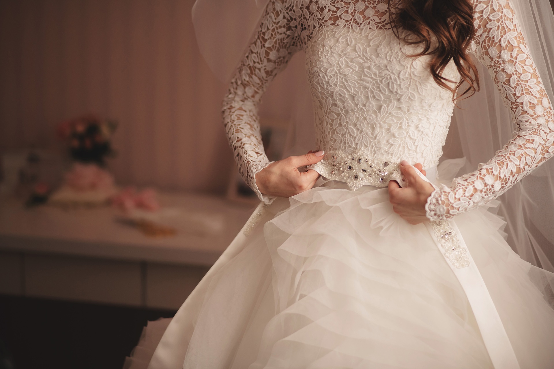 A bride in a white lace wedding dress adjusts her jeweled belt, with her face partially out of frame and a blurred bouquet in the background.