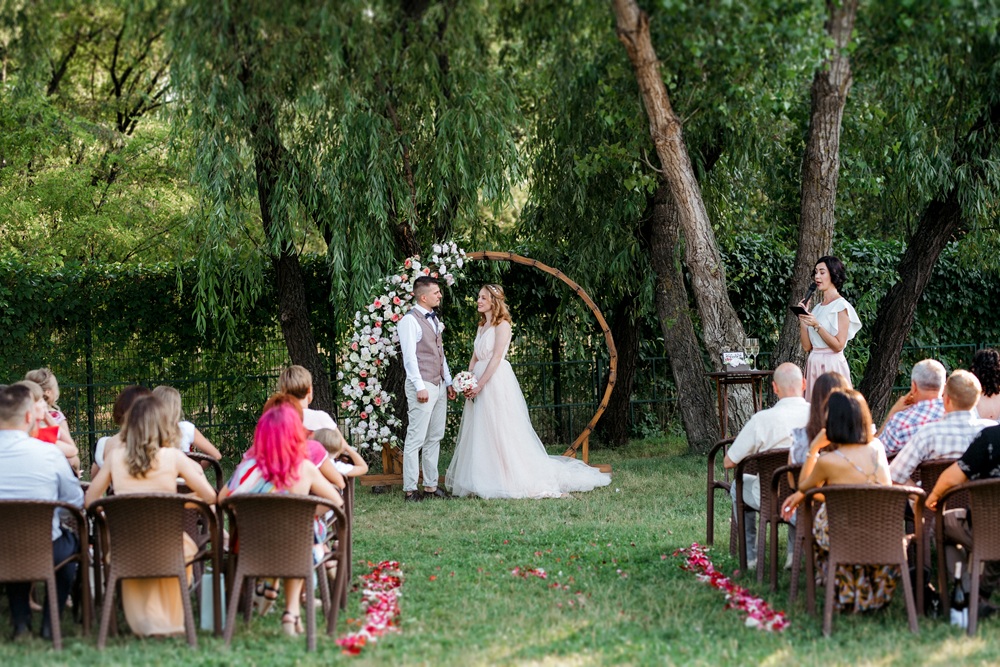 A bride and groom stand under a floral arch during an outdoor wedding ceremony, holding hands as guests sit in rows and an officiant reads from a book.