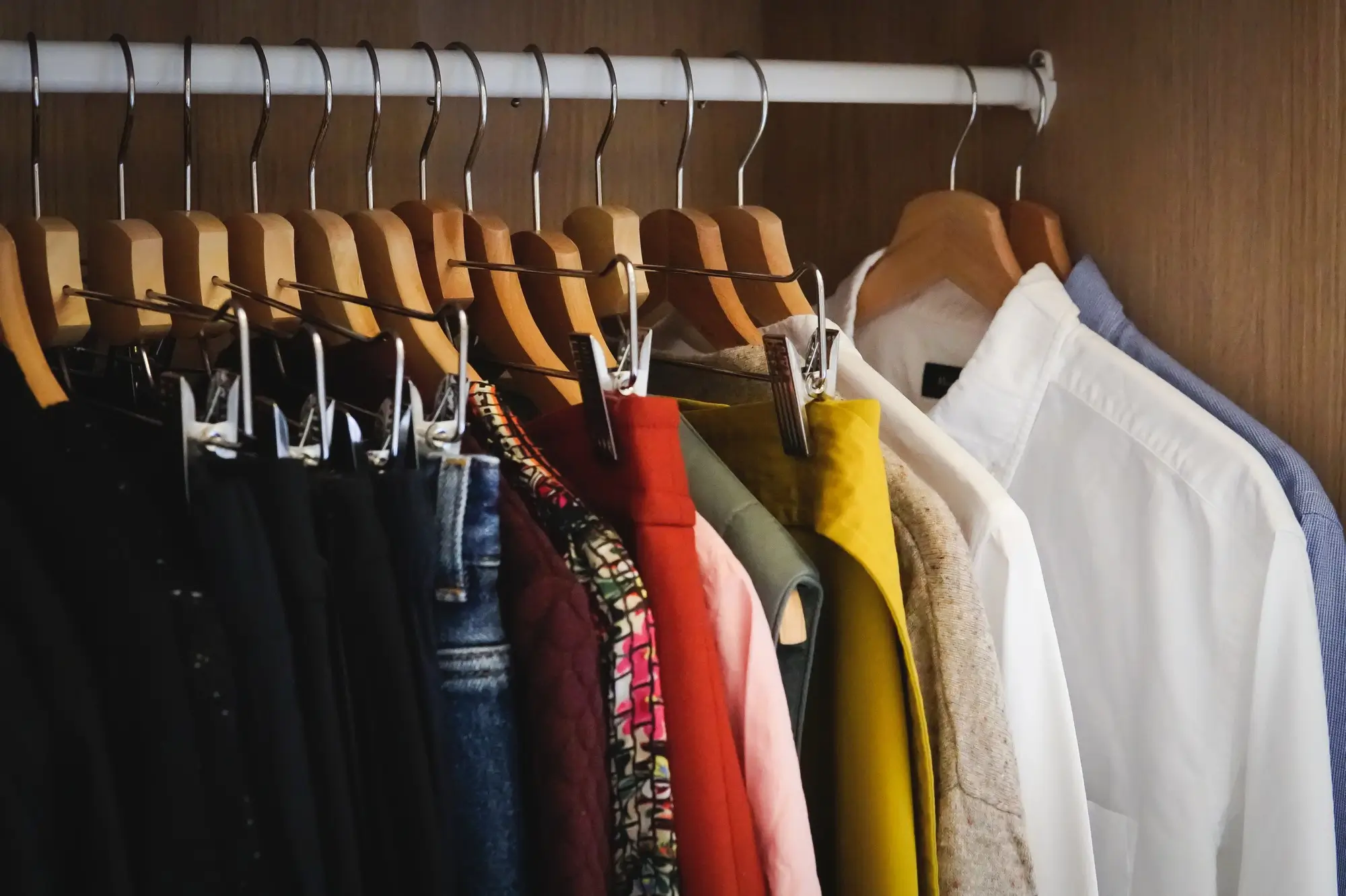 A row of assorted clothing, including shirts, jackets, and pants, hanging on wooden and metal hangers inside a closet.