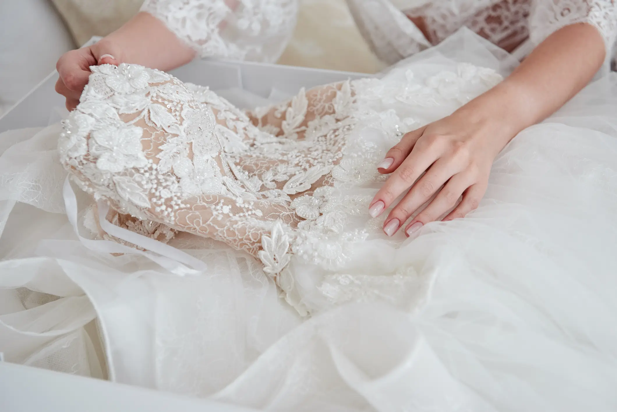 A person holds a white lace wedding dress with floral embroidery, laying it out on a soft surface.